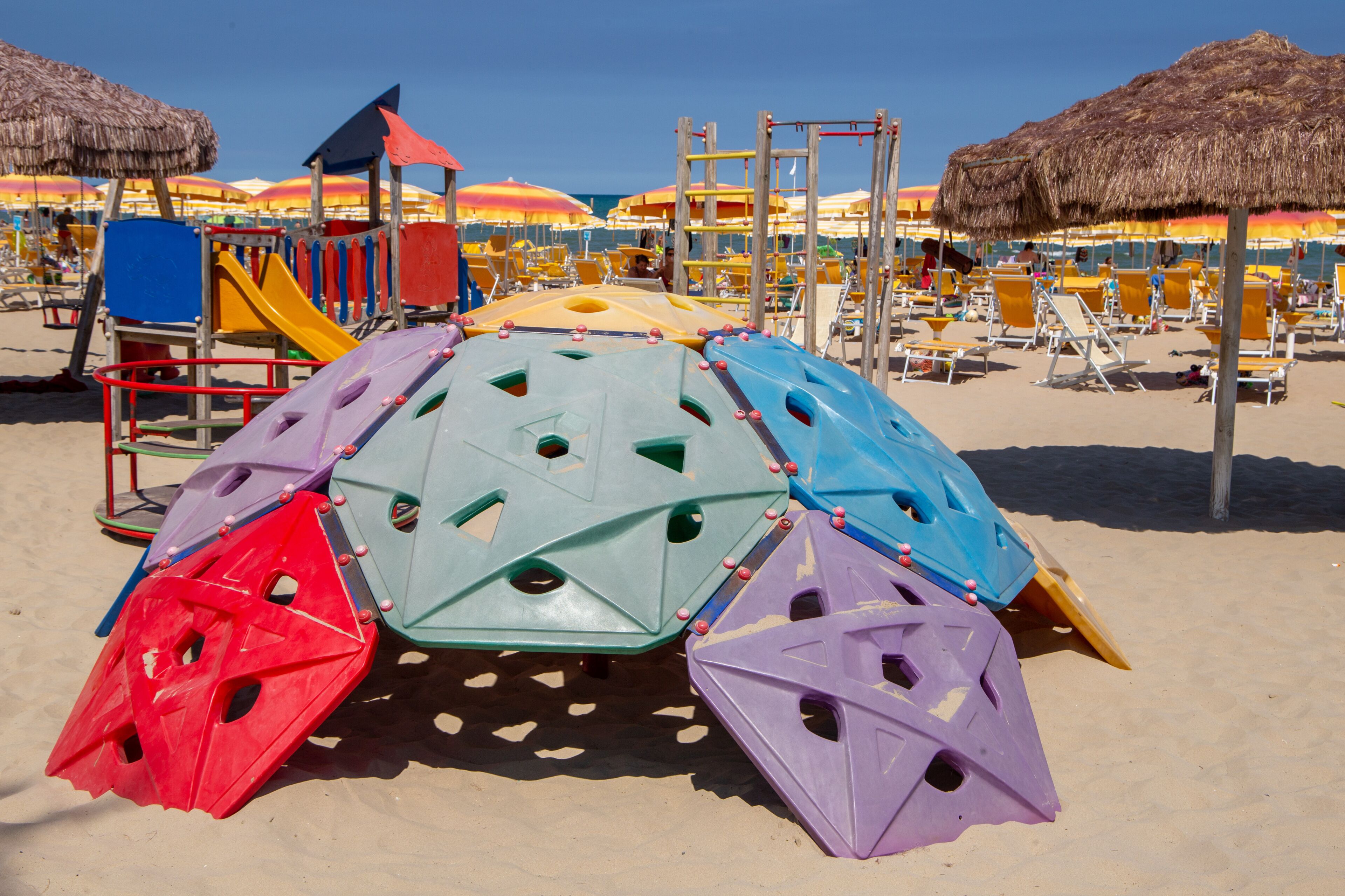 A beach play area with colorful climbing structures and thatched umbrellas.