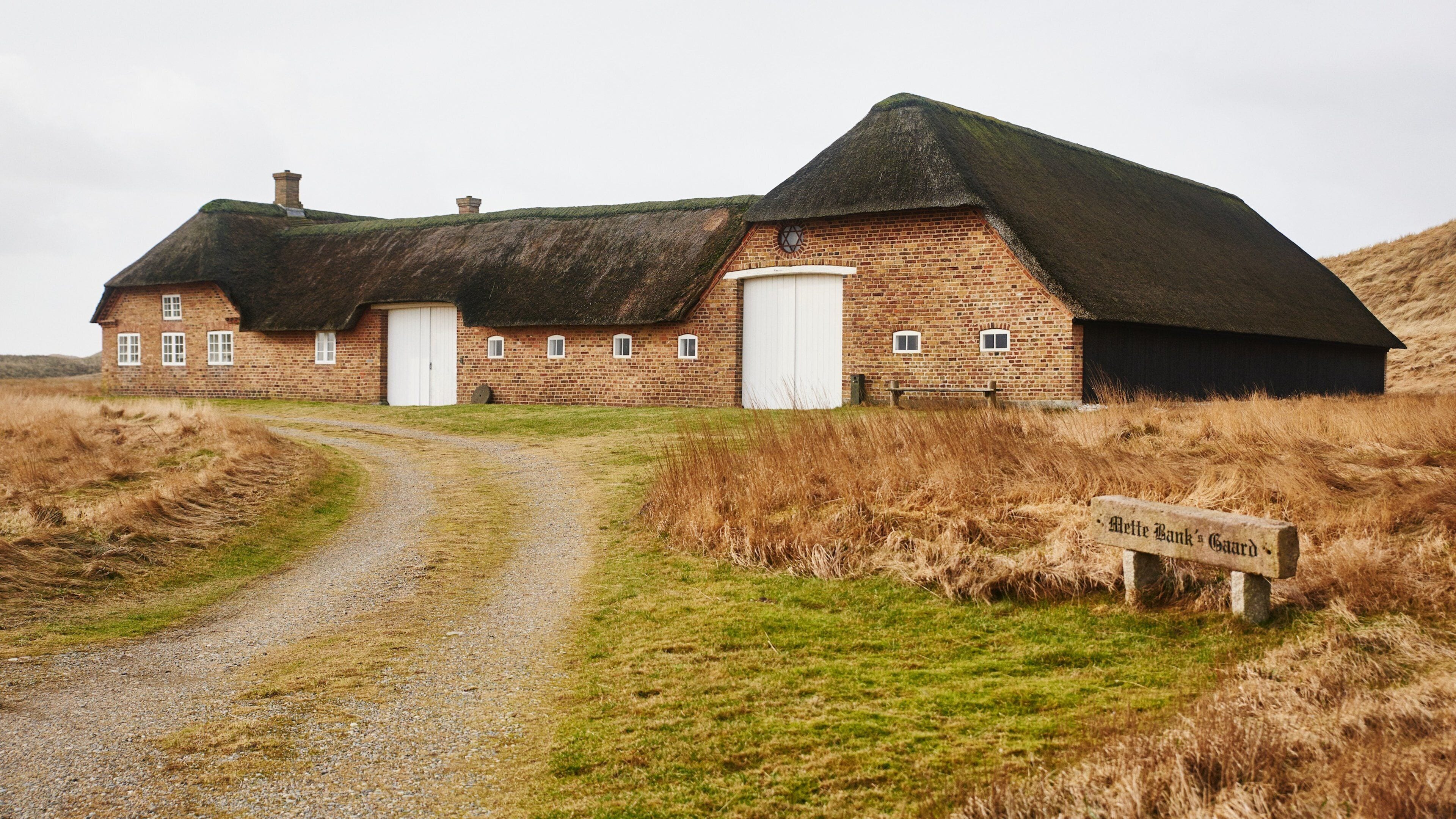 A traditional brick building with a thatched roof, a gravel path leading to it, and a sign reading "Alette Bank's Gaard".