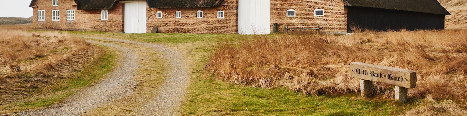A traditional brick building with a thatched roof, a gravel path leading to it, and a sign reading "Alette Bank's Gaard".