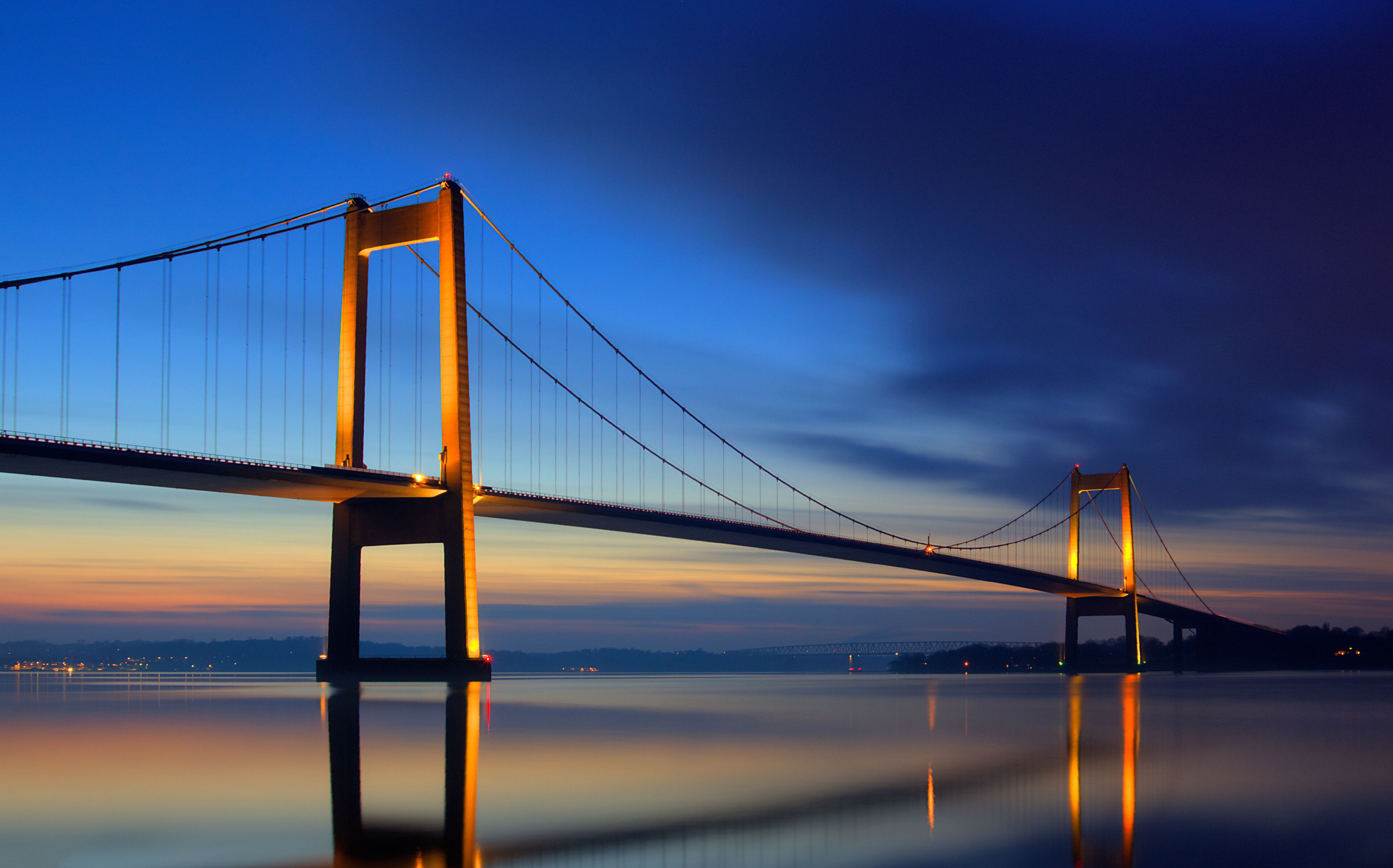 A suspension bridge at dusk with its reflection on calm water.