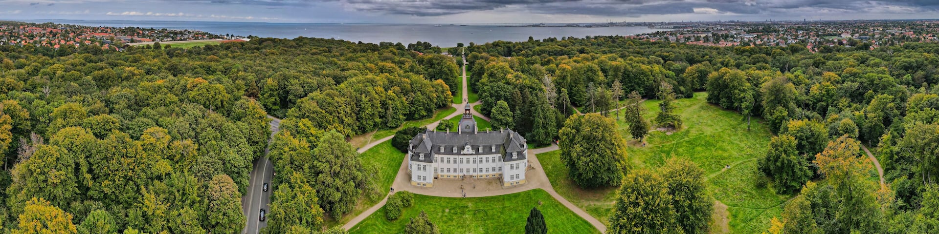 Aerial view of a grand, white palace nestled amidst lush green trees, leading the eye towards a serene sea under a dramatic, cloudy sky, Gentofte, Denmark.