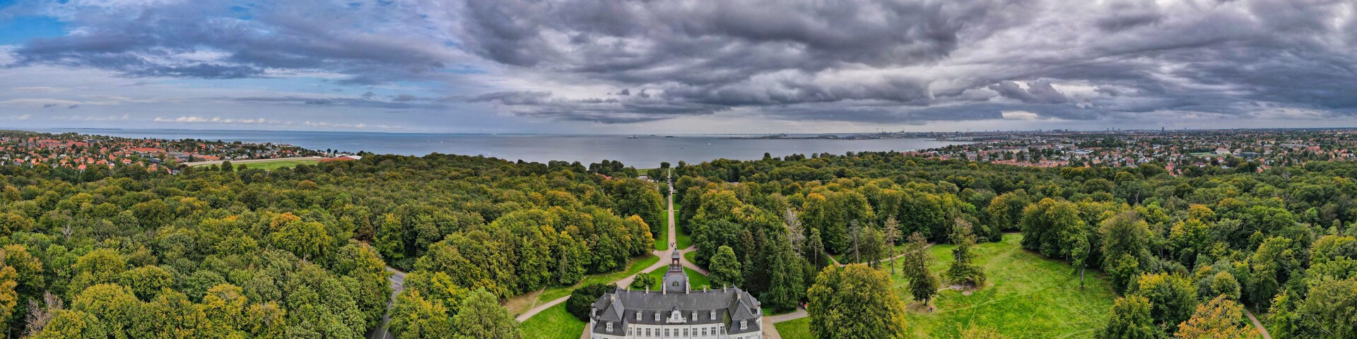 A large estate surrounded by dense forest, a lake, and a distant cityscape under a cloudy sky.