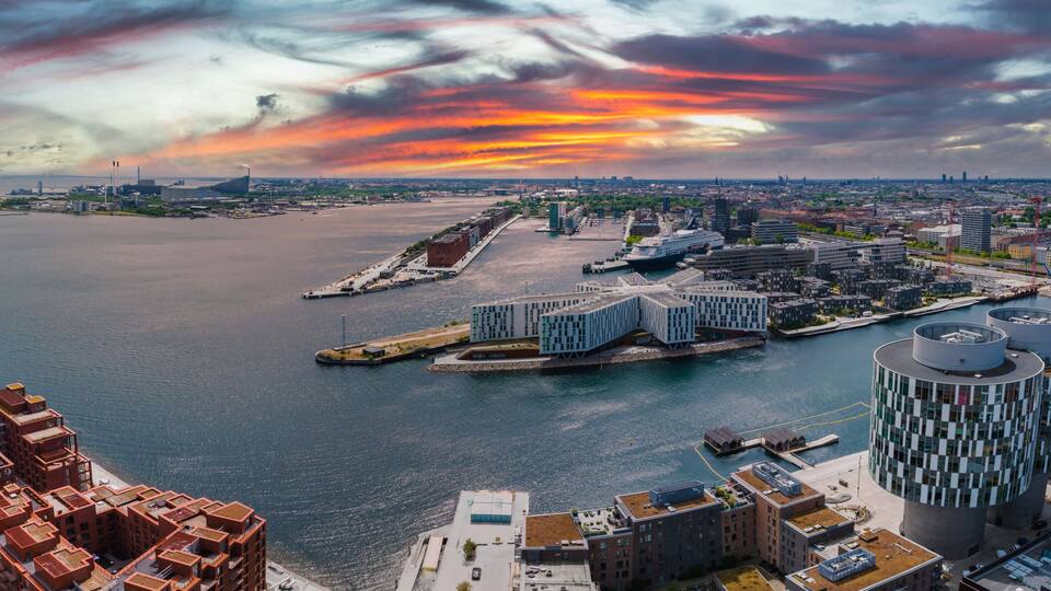 A waterfront cityscape with modern architecture, a large body of water, and a dramatic sunset sky.