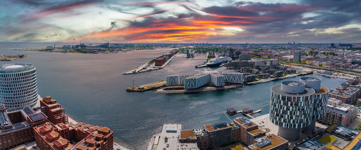 A waterfront cityscape with modern architecture, a large body of water, and a dramatic sunset sky.