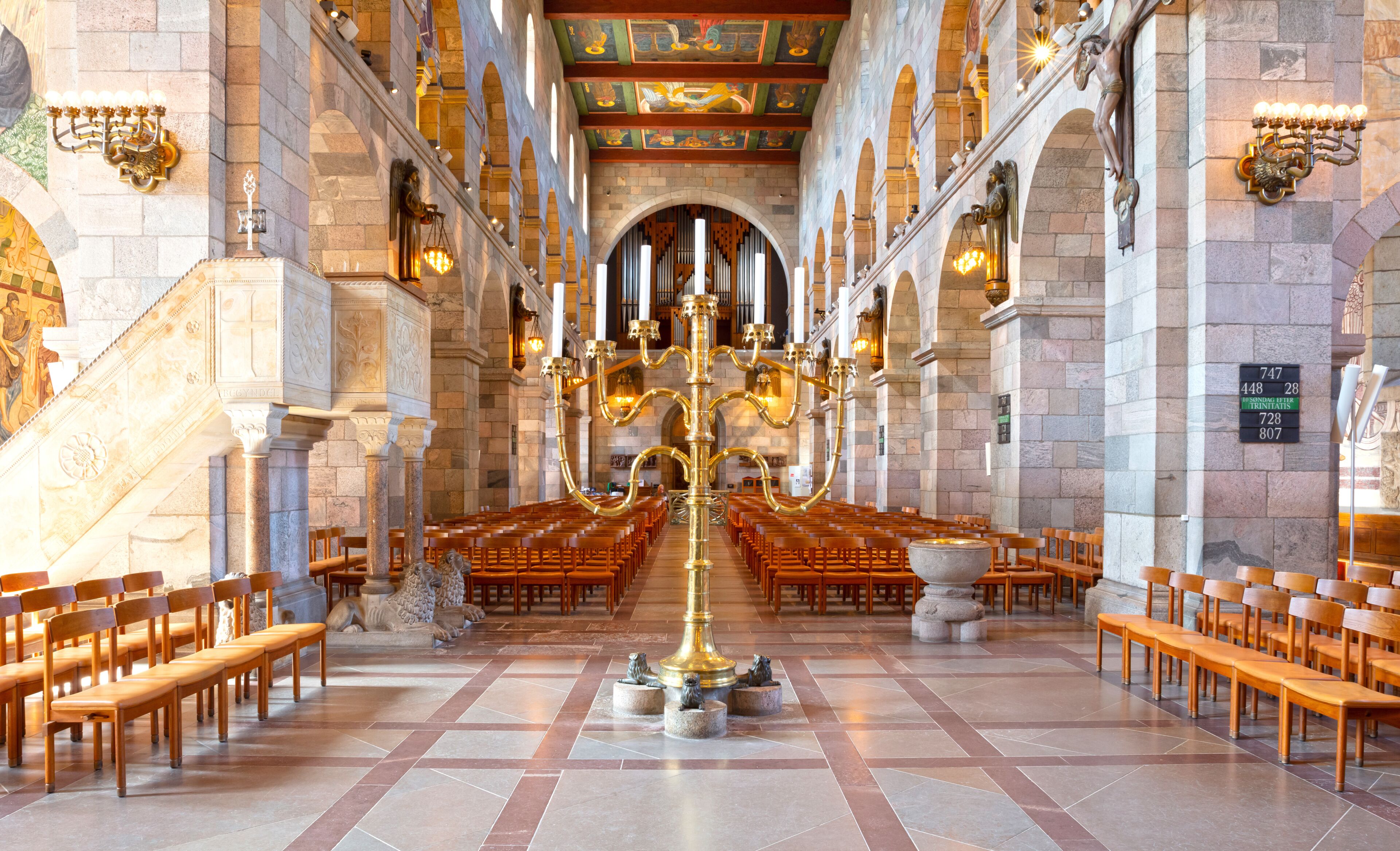 A church interior with rows of wooden pews, a central golden candelabrum, and stone columns.