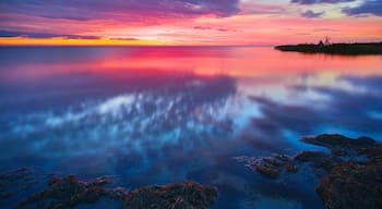 A sunset over a calm sea with a rocky shoreline and a distant island with a windmill.