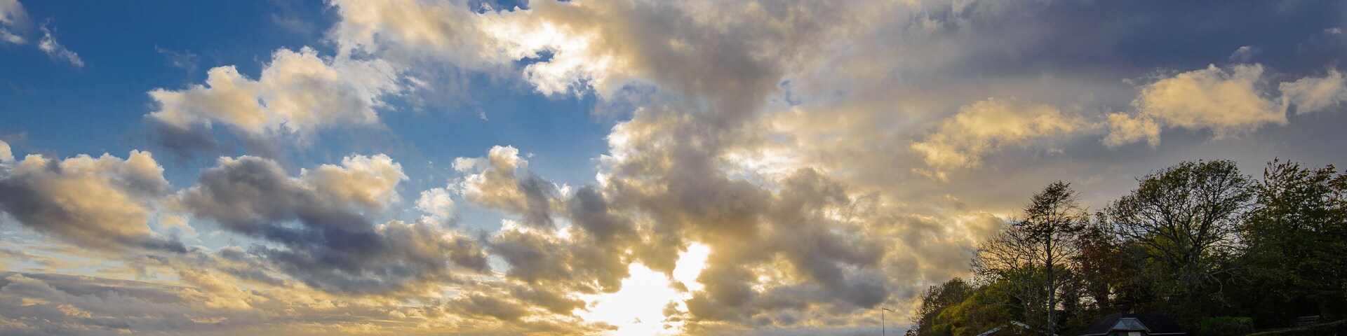 Paisagem costeira com pedras grandes em primeiro plano, um corpo d'água e um pôr do sol com nuvens dispersas.