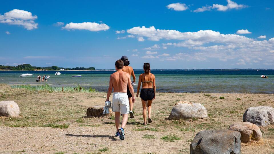 A beach with people walking, large rocks, and boats in the water.