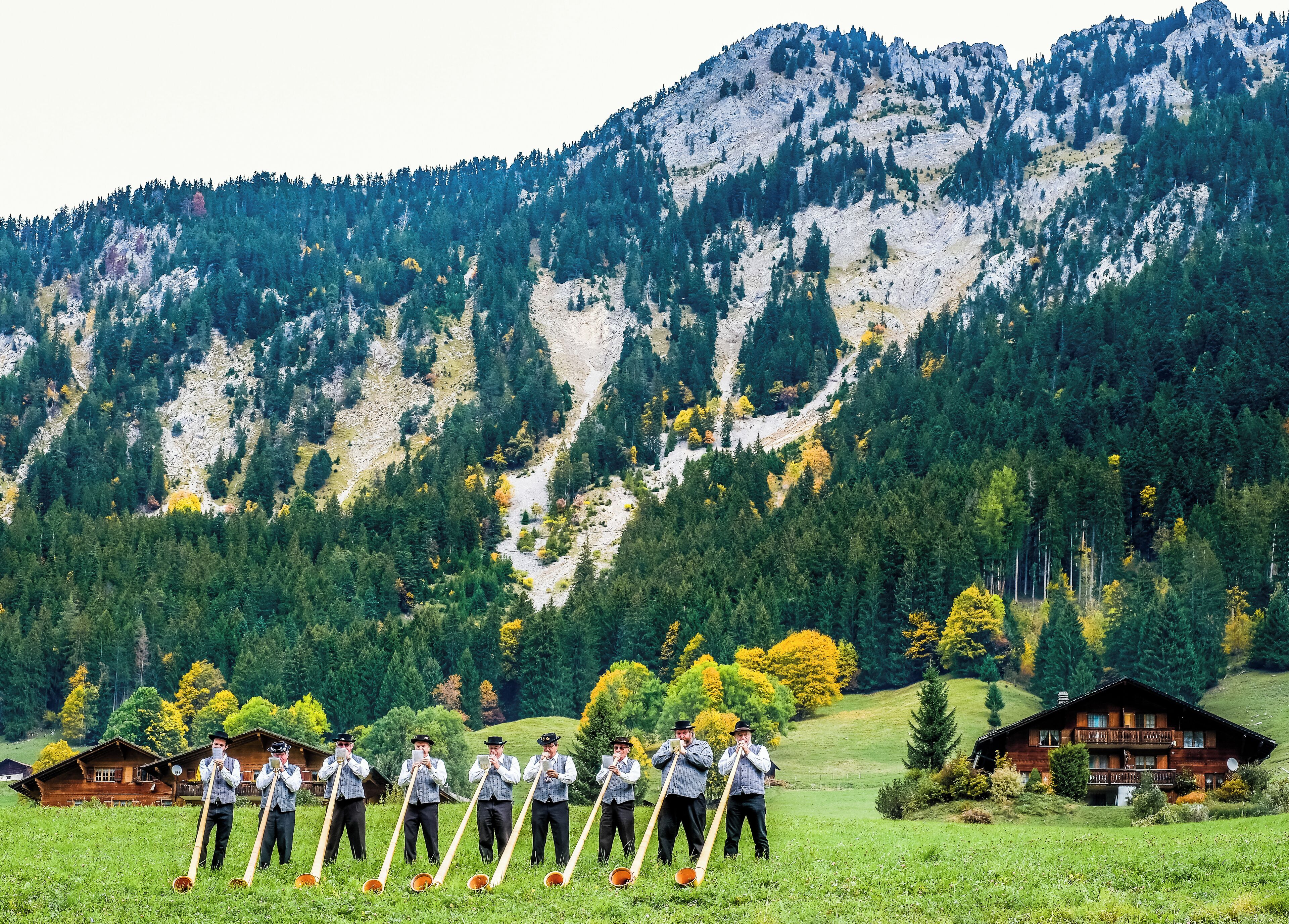 A group of men in traditional attire holding wooden spoons in a grassy field with a mountain backdrop.