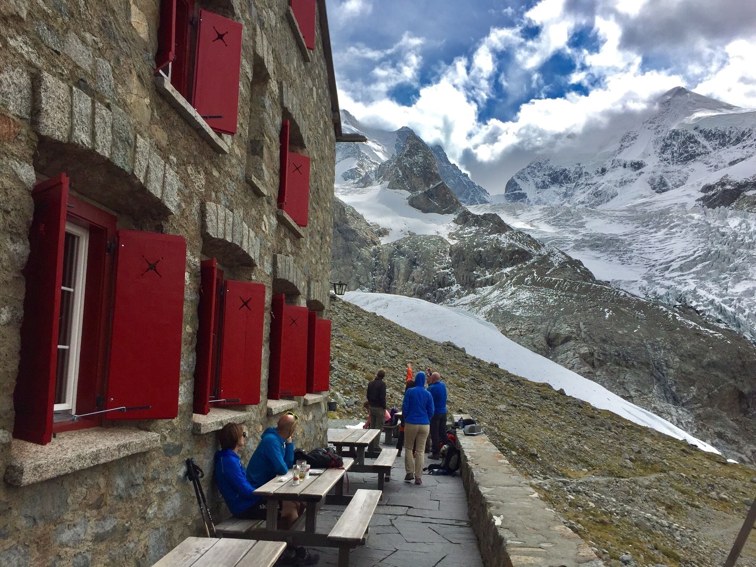 A stone building with red shutters and a view of a snowy mountain range.