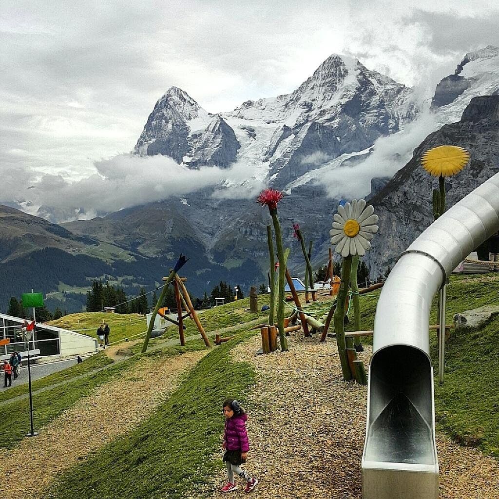 Un parque infantil con tobogán y estructuras para escalar, con montañas nevadas y un cielo nublado al fondo.