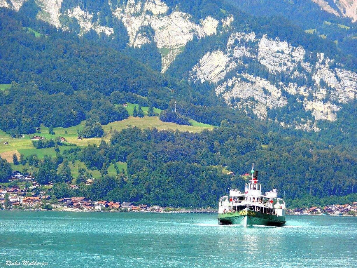 A boat on a turquoise lake with a mountainous backdrop and a village on the shore.