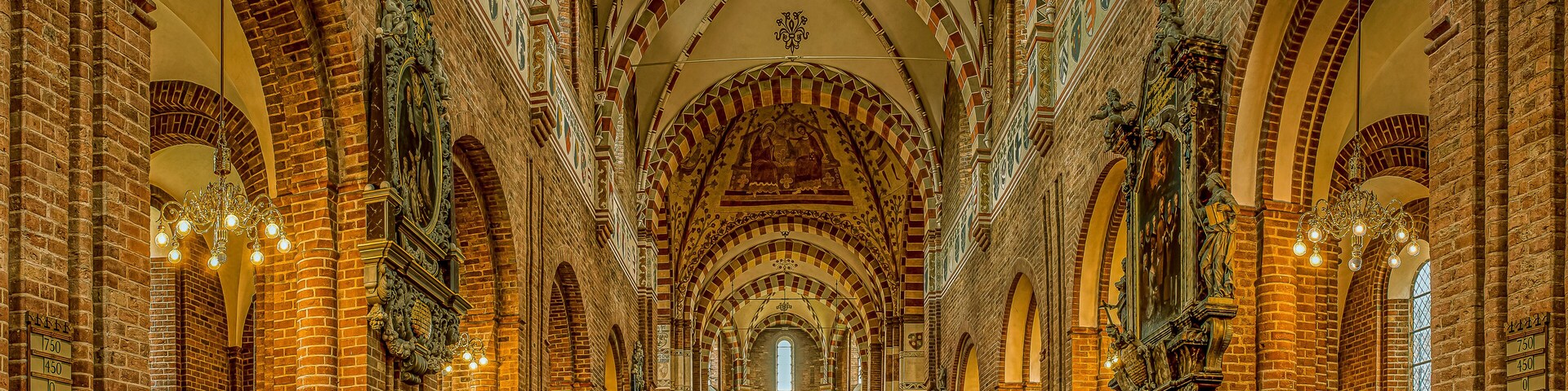 Interior de uma catedral com arcos de tijolo, uma nave central e lustres elaborados.