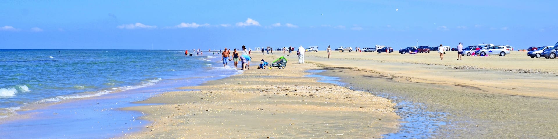A beach with people walking, parked cars, and a calm sea.