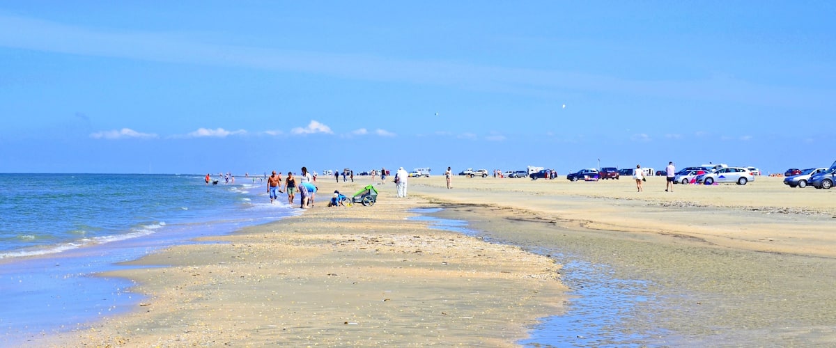 A beach with people walking, parked cars, and a calm sea.