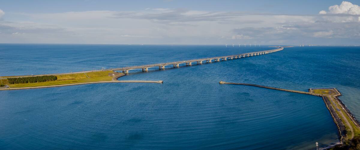 A long bridge spanning a wide body of water, with a curved shoreline and a breakwater on the right.