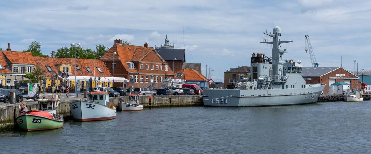 A harbor with a large naval ship, smaller boats, and traditional buildings along the waterfront.
