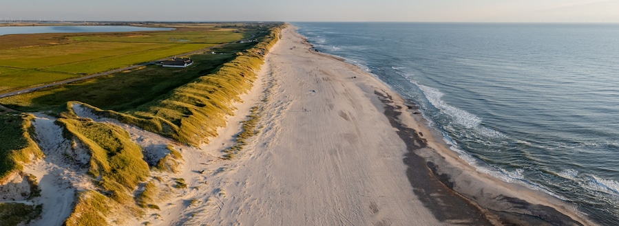 Aerial view of long, untouched coastline with lush green meadows, golden dune and sandy beach meeting sea with gentle waves on one side while tranquil waters of Ringkøbing Fjord stretch on other side