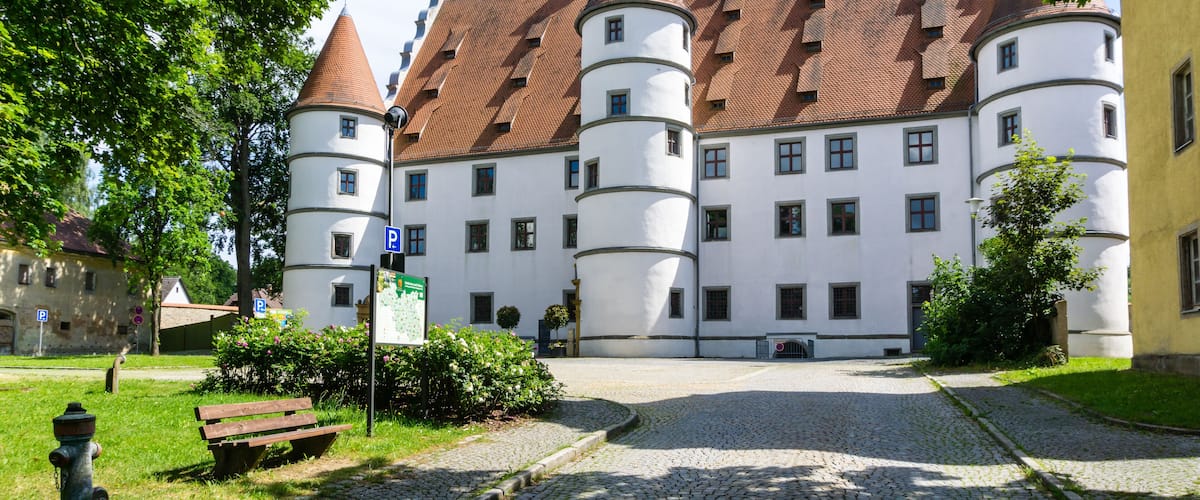 A historic building with white walls and red-tiled roofs, featuring multiple towers and a cobblestone courtyard.
