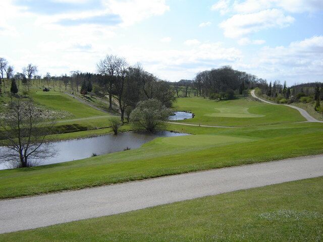 A golf course with a pond, a walking path, and a wooded area.