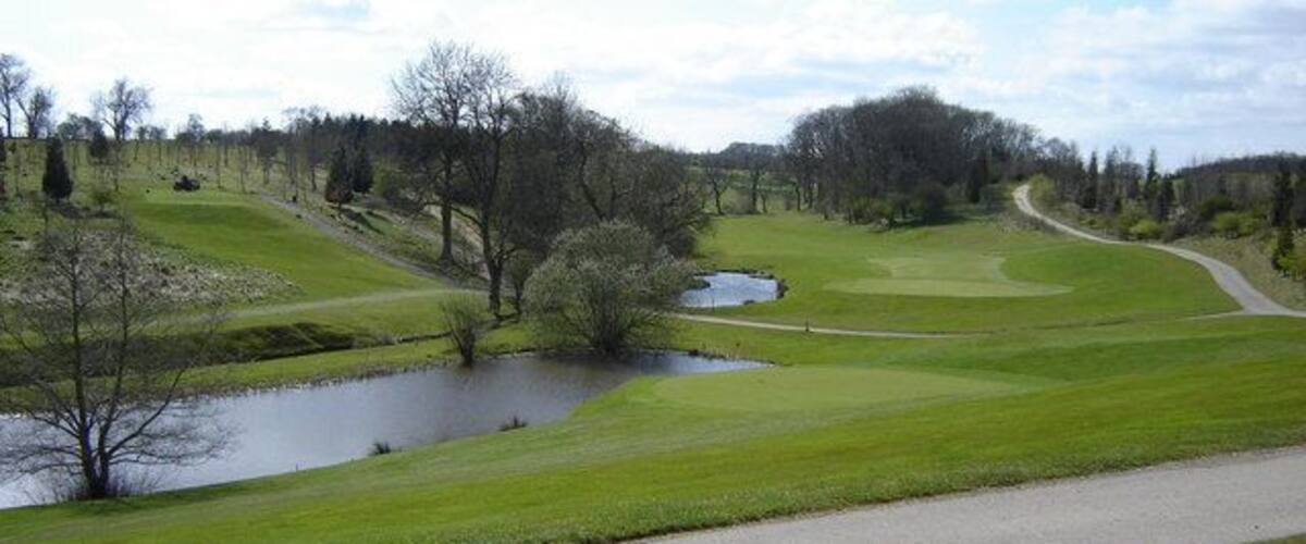 A golf course with a pond, a walking path, and a wooded area.