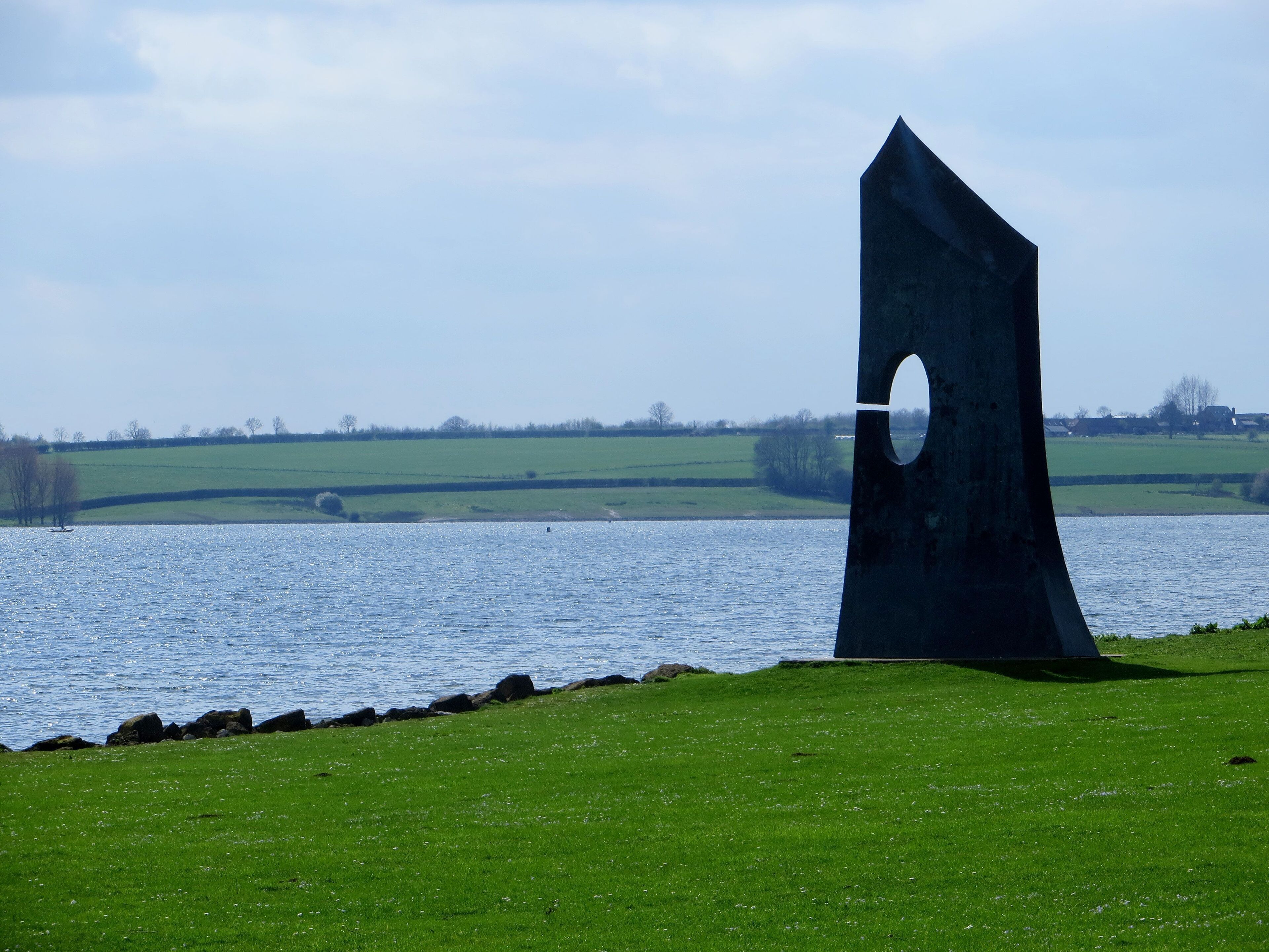 A large, dark sculpture in a grassy field by a body of water.