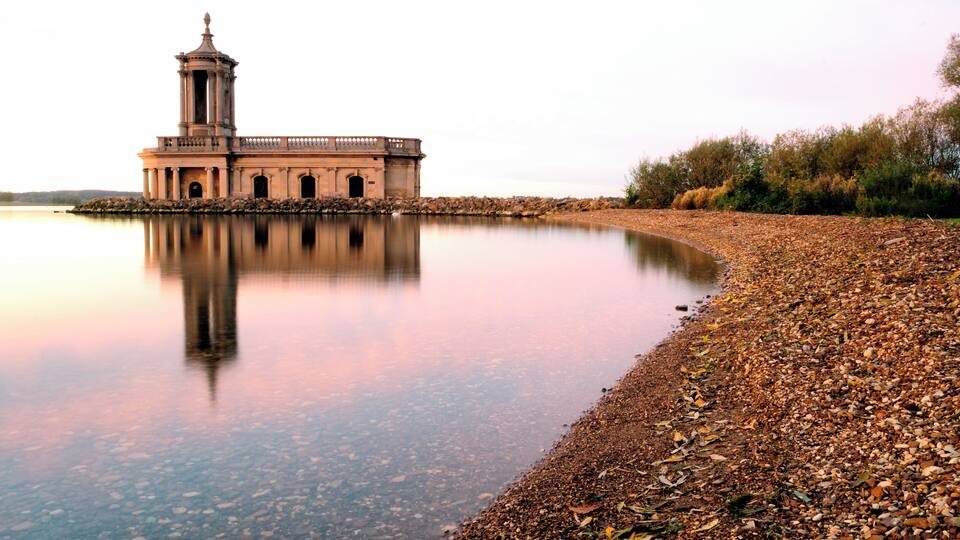 A historic building with a tower by a calm lake with a pebble beach.