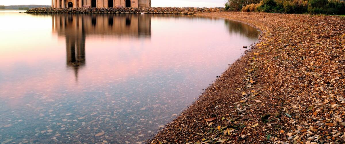 A historic building with a tower by a calm lake with a pebble beach.