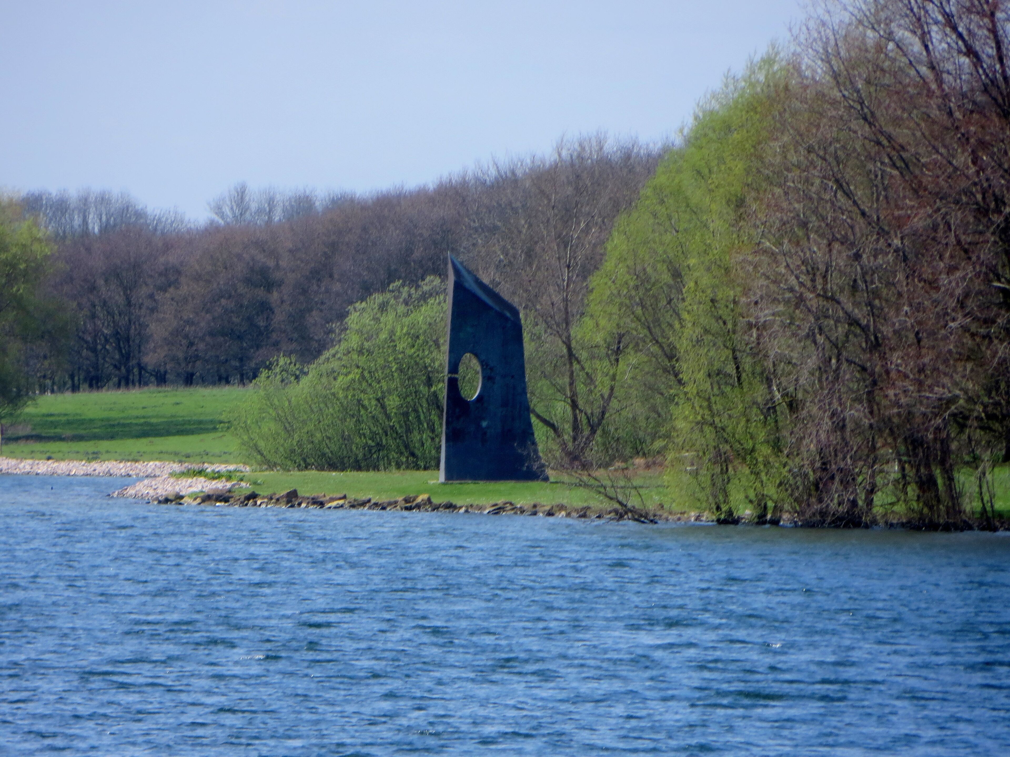 A sculpture by the water, surrounded by trees and a grassy area.