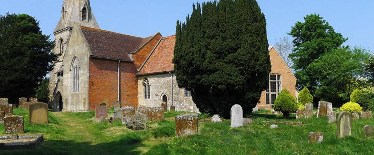 A church with a tall steeple, surrounded by a cemetery with headstones and a large tree.
