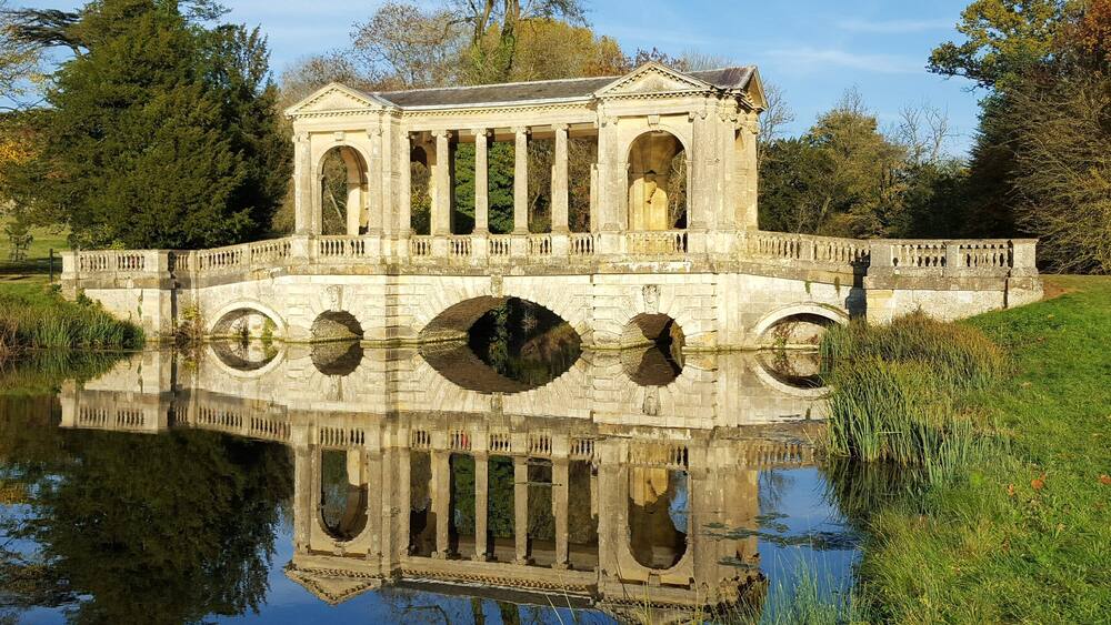 A stone bridge with arches over a calm body of water, surrounded by trees and grass.