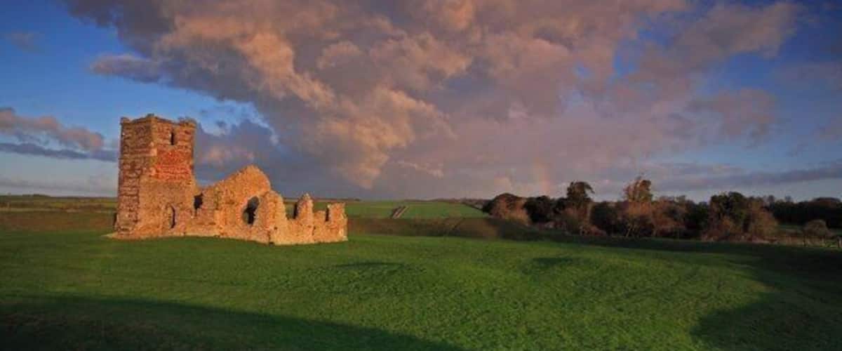 Les vestiges d’un ancien bâtiment en pierre, avec une tour, se dressent sur un champ verdoyant, le tout sur fond d’un ciel spectaculaire.