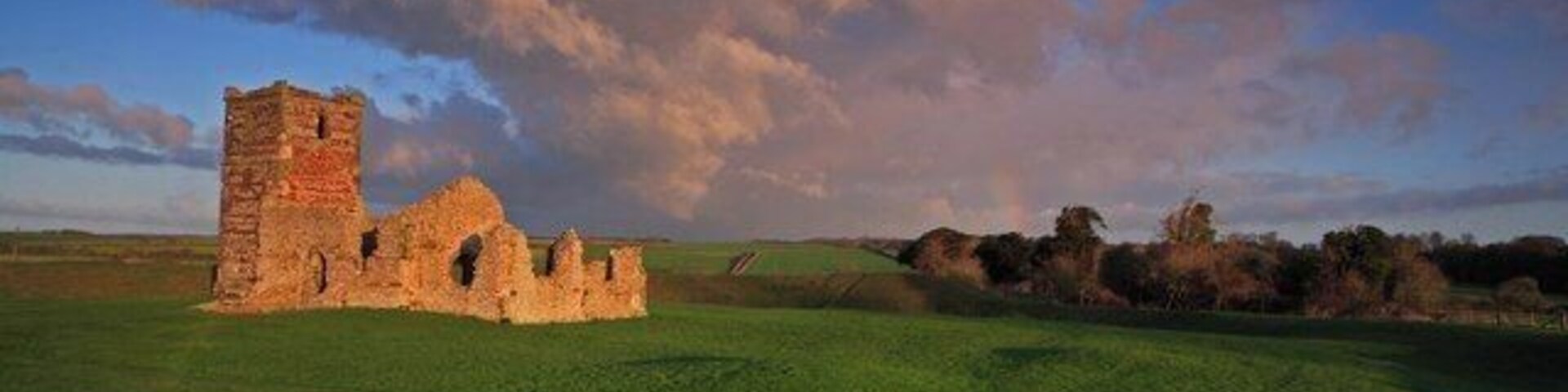 Ruinas de piedra históricas con una torre, frente a un campo cubierto de hierba y un cielo impresionante.