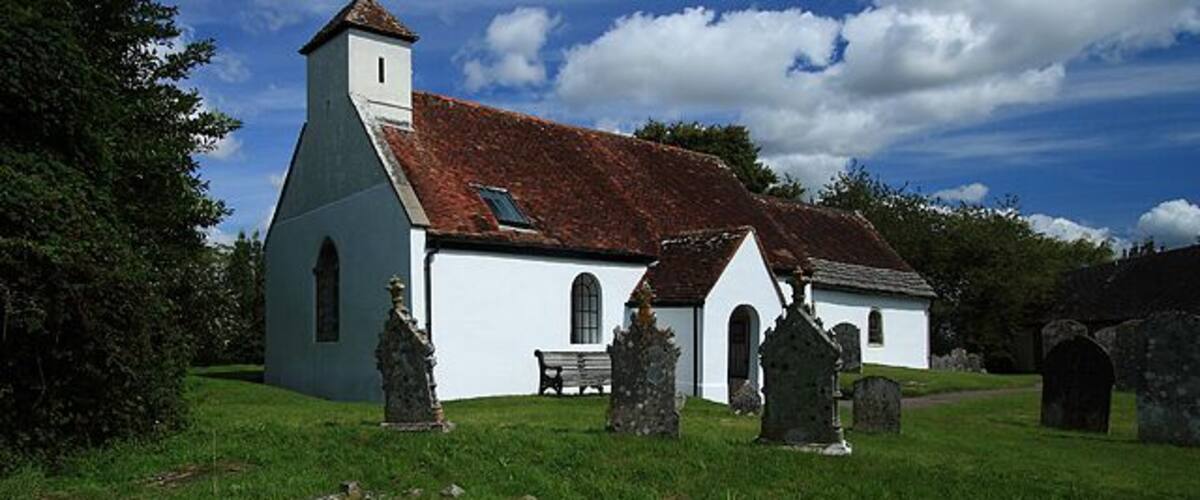 A white church with a red roof, a stone bench, and several gravestones in a grassy field.
