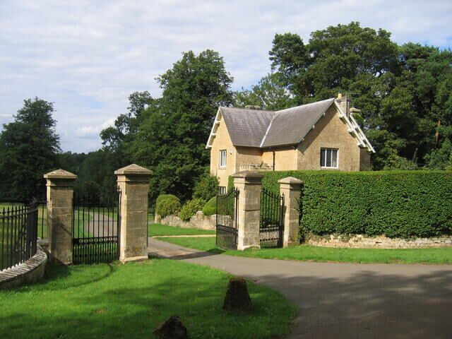 A stone house with a grey roof, surrounded by a hedge and trees.