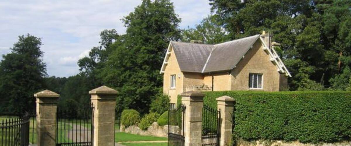 A stone house with a grey roof, surrounded by a hedge and trees.