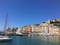 Une ville côtière aux bâtiments aux couleurs vives, un port de plaisance rempli de bateaux et une église imposante située sur une colline.