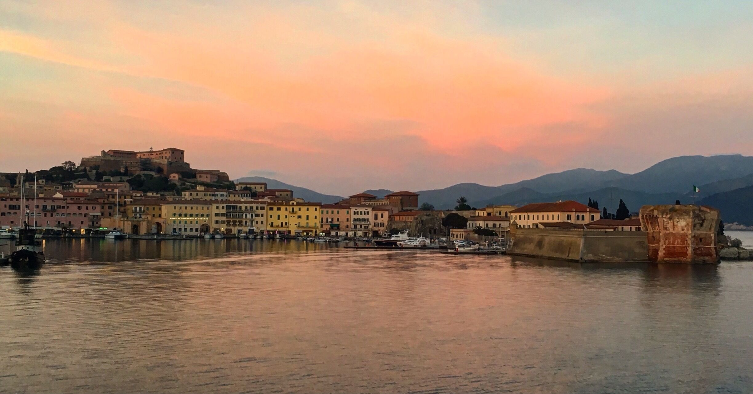 A coastal cityscape with a prominent castle on a hill, a harbor with boats, and a backdrop of mountains.
