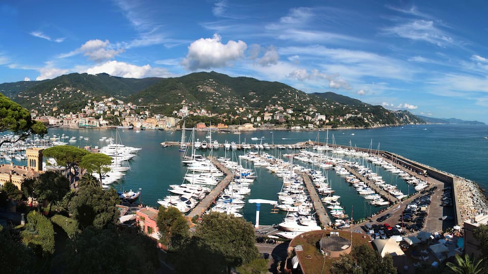 A marina with numerous boats docked, surrounded by lush green hills and a clear blue sky.