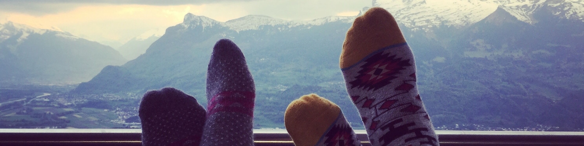 Pés com meias coloridas apreciando uma paisagem montanhosa com picos nevados e uma pequena aldeia lá embaixo.