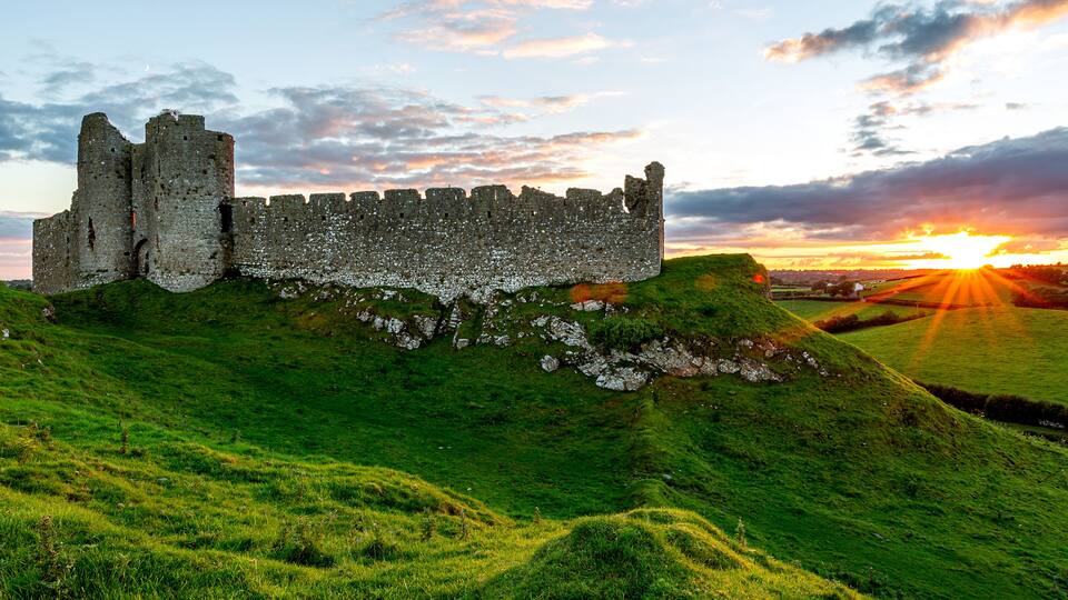 A historic stone castle on a grassy hill with a sunset in the background.