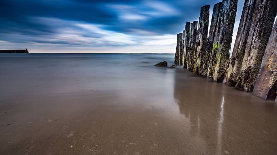 A beach with a long row of weathered wooden posts and a calm sea under a cloudy sky.