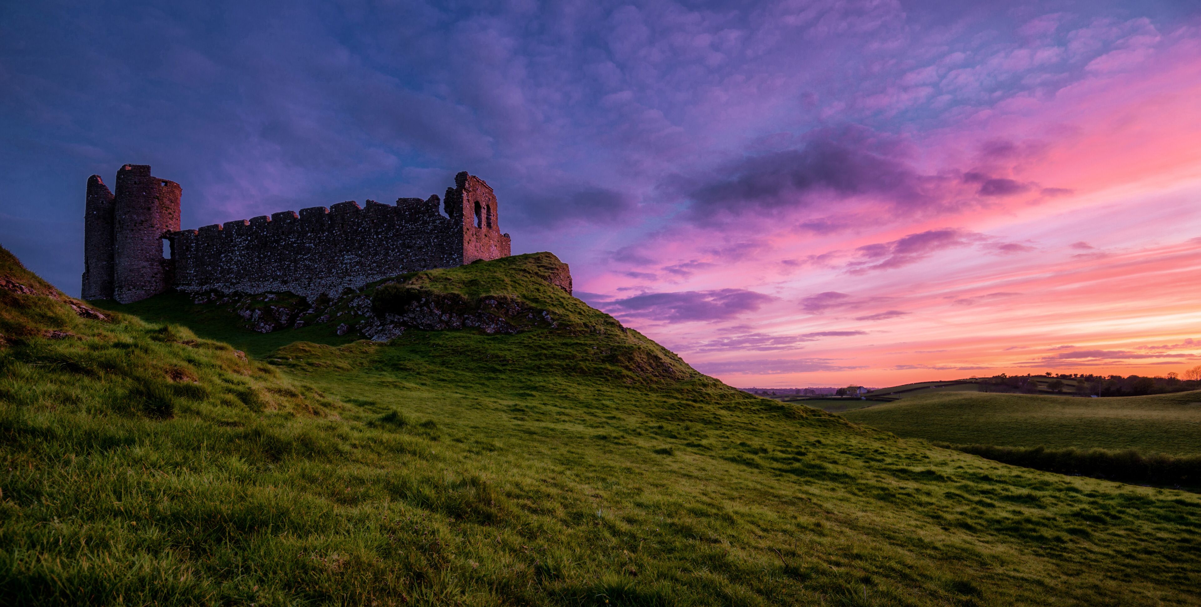 A historic castle on a grassy hill with a sunset sky.