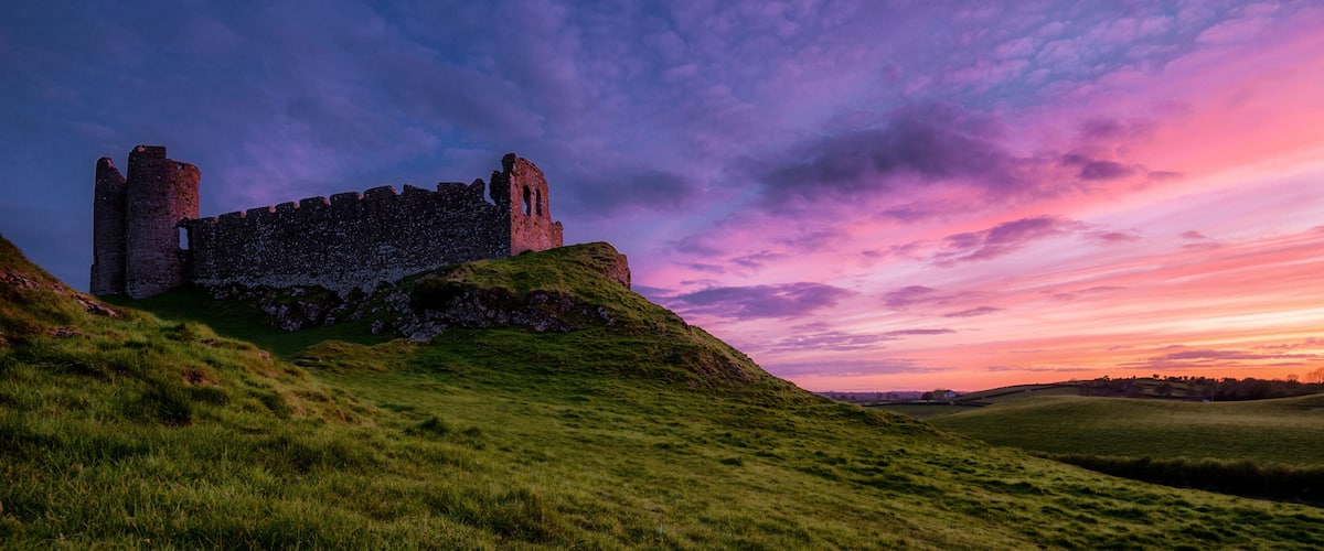 A historic castle on a grassy hill with a sunset sky.