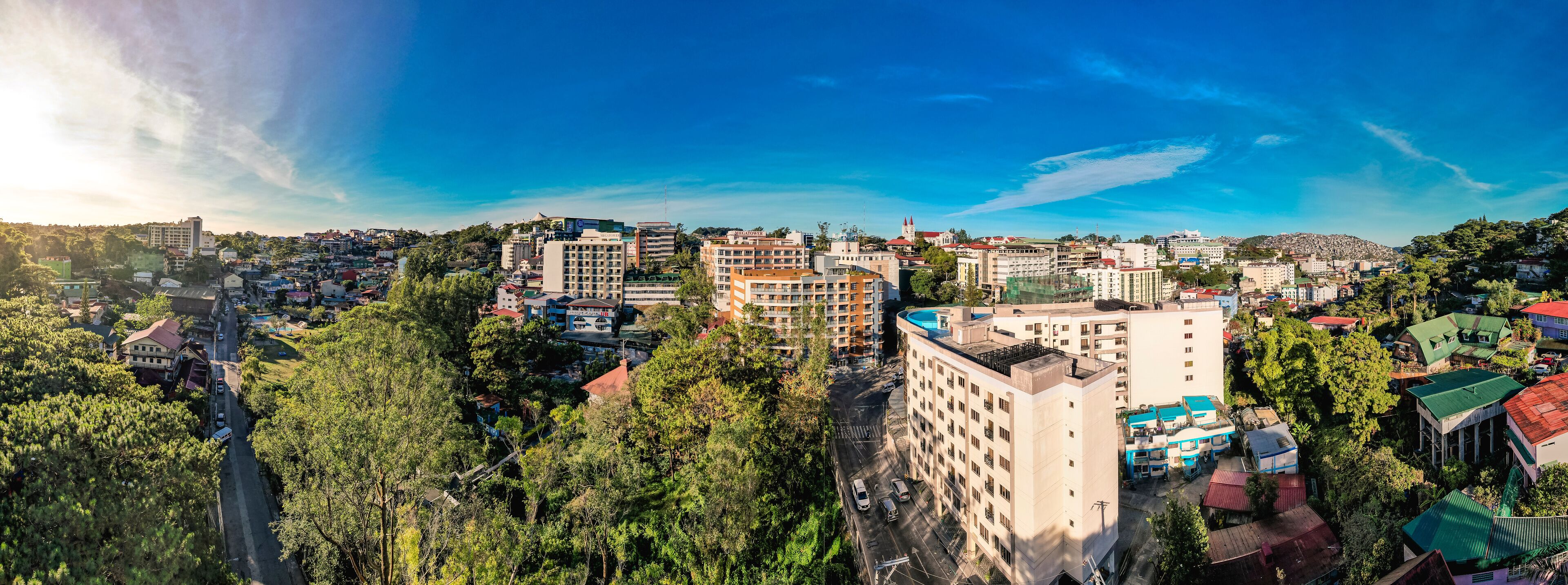 A cityscape with a mix of residential and commercial buildings, surrounded by lush greenery and a clear blue sky.