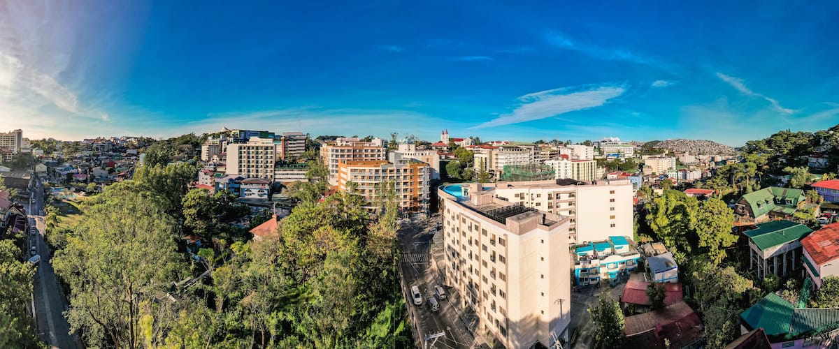 A cityscape with a mix of residential and commercial buildings, surrounded by lush greenery and a clear blue sky.