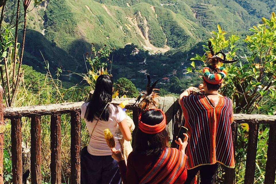 Um grupo de pessoas em um caminho de pedra, com vista para um vale com colinas verdes e uma estrada sinuosa.