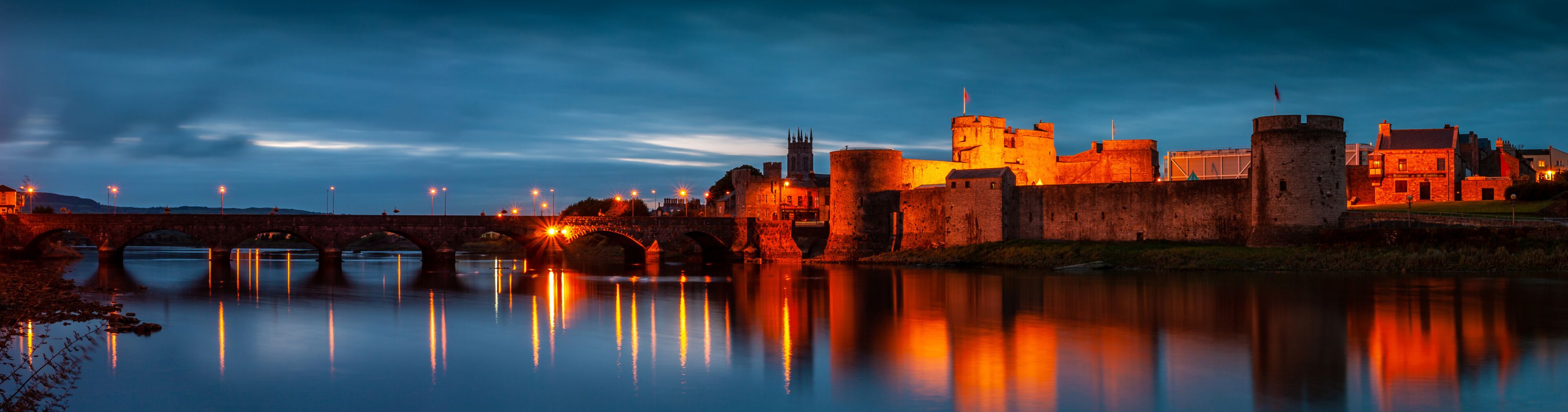 A historic castle with a moat and a bridge, illuminated at dusk.
