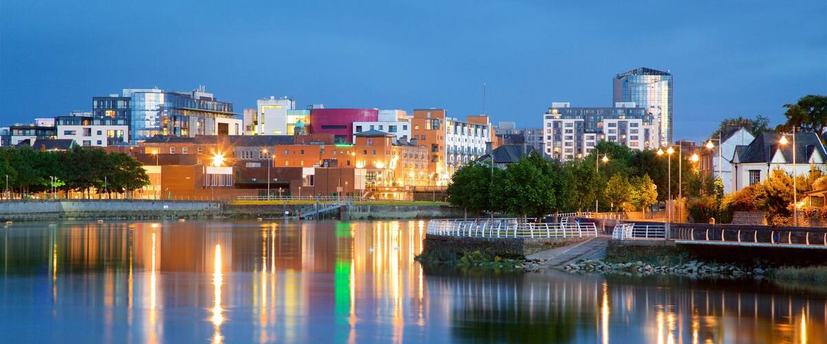 A city skyline at dusk with illuminated buildings reflecting on the water.