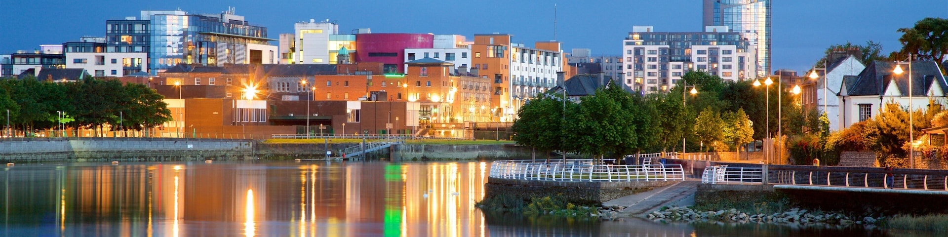 A city skyline at dusk with illuminated buildings reflecting on the water.
